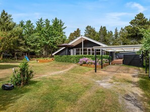 Una casa moderna con un porche cubierto, un área de juegos con tobogán y un jardín con plantas florecientes.