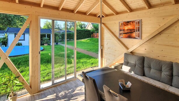 A wooden interior with a dining area, a sofa, and a large window offering a view of a garden and neighboring houses. - Ferienwohnung Neppermin/Usedom (Neppermin/Usedom)