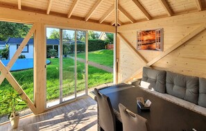 A wooden interior with a dining area, a sofa, and a large window offering a view of a garden and neighboring houses.