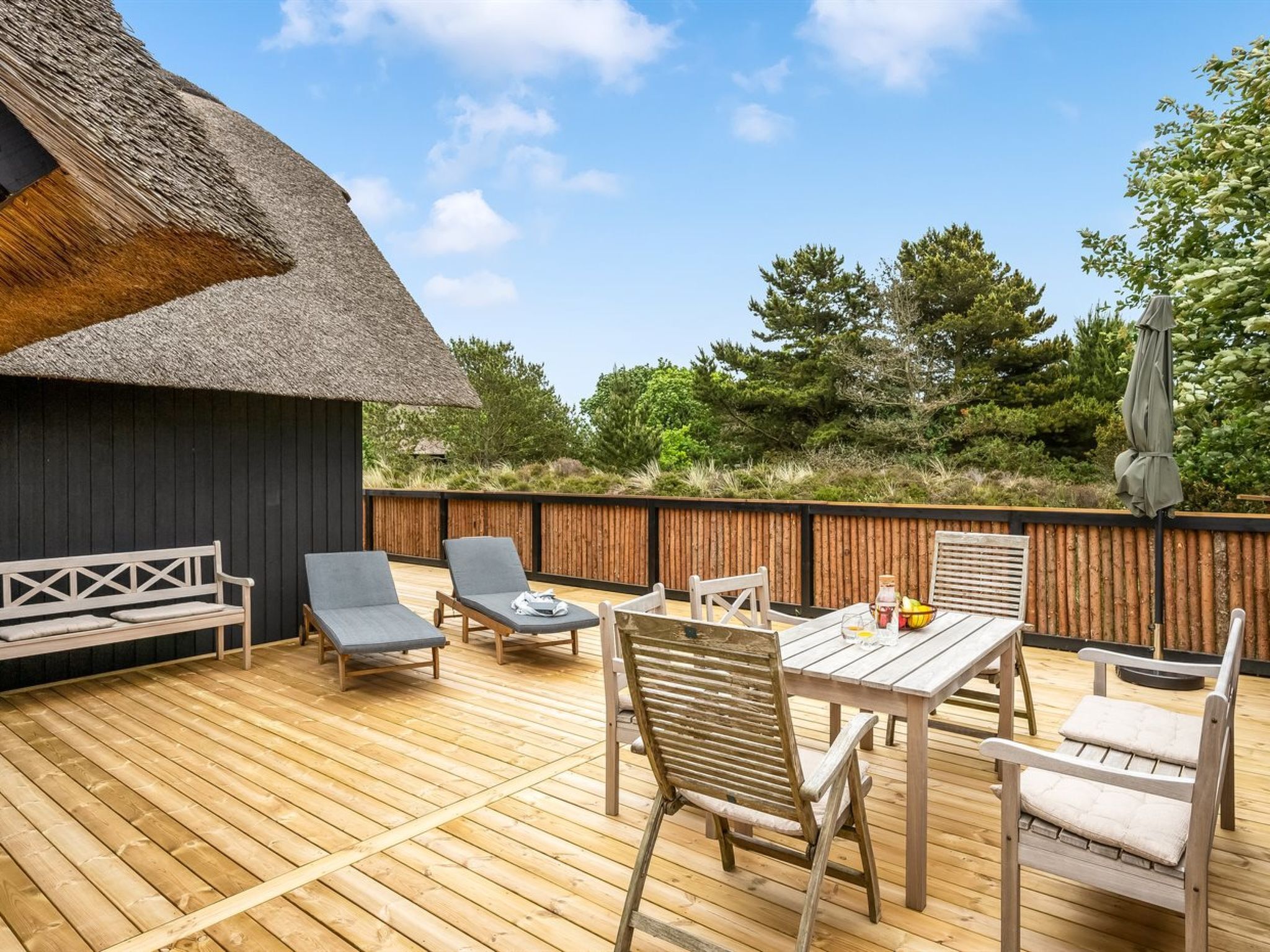 Eine Holzterrasse mit Tisch und Stühlen, ein Gebäude mit Strohdach und ein Holenzaun.
