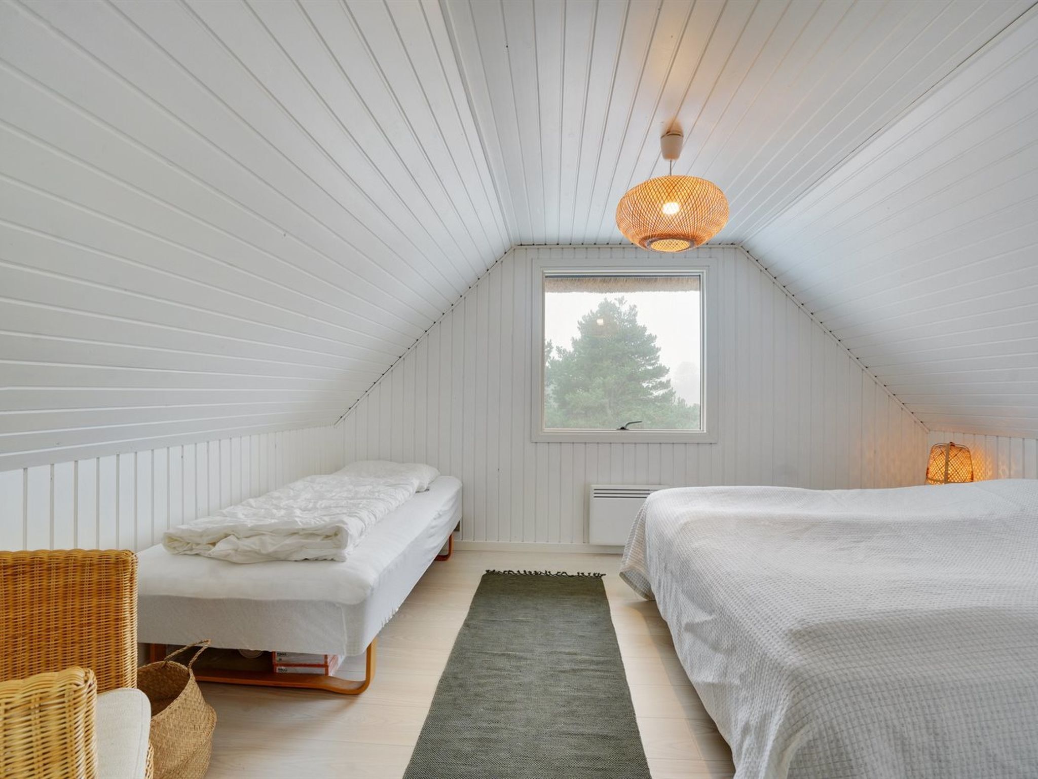 Un dormitorio bajo de techo, pequeño y con paneles blancos, con dos camas, una ventana, una lámpara colgante y una cesta tejida.