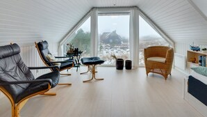 Een ruime zolderkamer met schuine wanden, een groot raam met uitzicht op een huis in de verte, en een knus zitgedeelte met lederen en rieten stoelen.