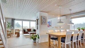 Een ruime woonkamer met een houten eettafel en stoelen, een bank en een groot raam met uitzicht op de omgeving.