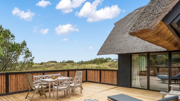 Een houten terras met een tafel en stoelen, een loungehoek met ligbedden, en een constructie met rieten dak en glazen deuren.