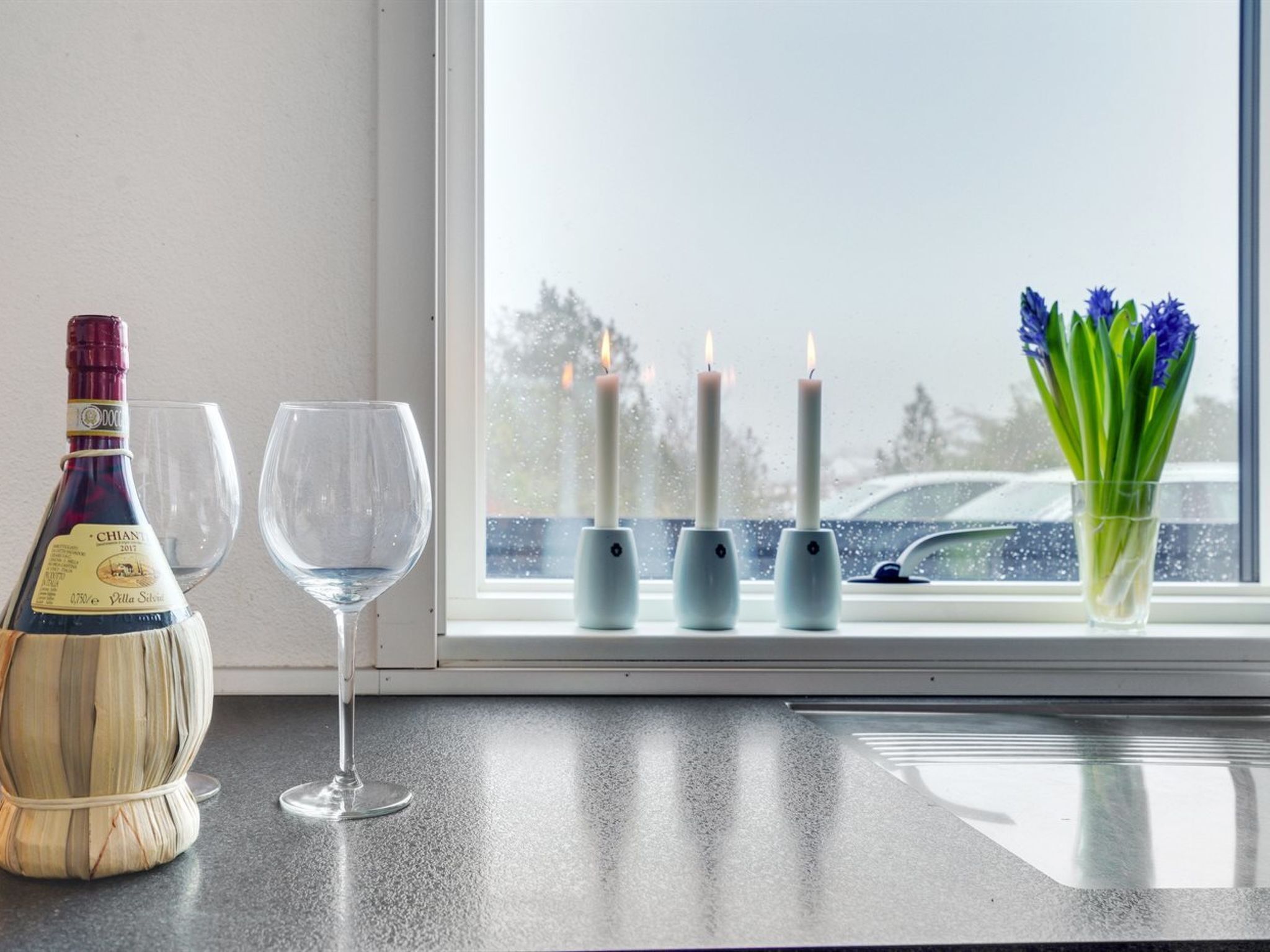 A bottle of Chianti wine, two wine glasses, and a vase with flowers on a window sill.