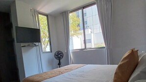 Et hotelværelse med en seng, et fladskærms-tv på væggen, en standventilator og et vindue med gardiner.