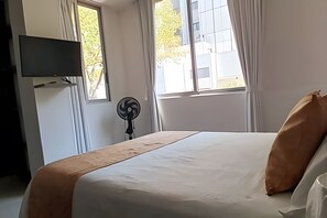 Ein Hotelzimmer mit einem Bett, einem an der Wand montierten Fernseher, einem Standventilator und Fenstern mit Vorhängen.