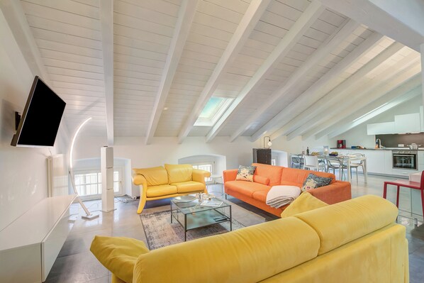 A modern living room with a high ceiling, a flat-screen TV mounted on the wall, and a kitchen area in the background.