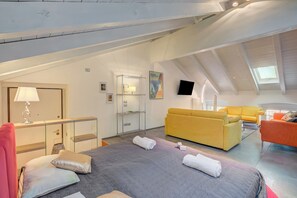 A spacious loft bedroom with a large bed, a yellow sofa, an orange sofa, a desk with a lamp, and a TV mounted on the wall.