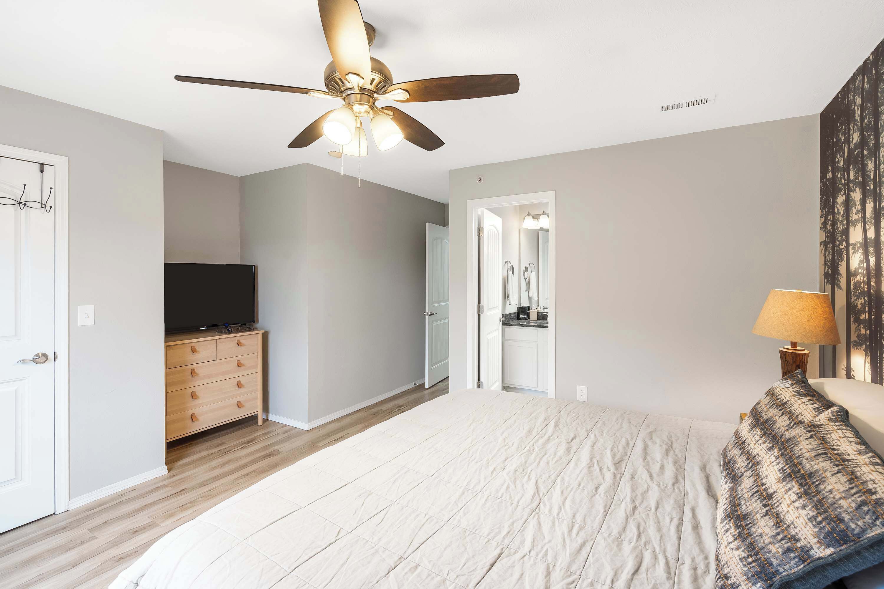 A bedroom with a bed, a ceiling fan, a dresser, a TV, and a bathroom visible through an open door.