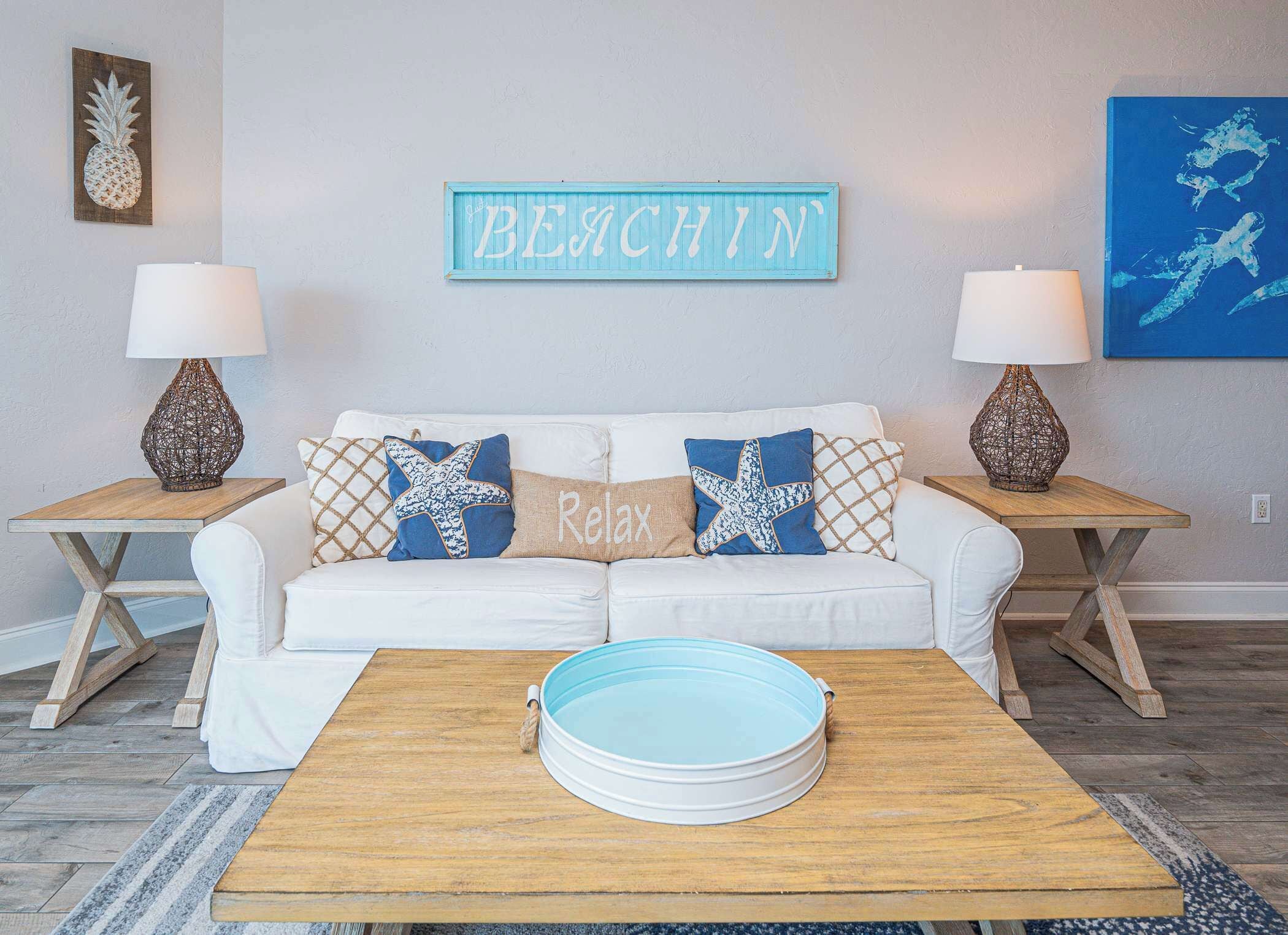 A living room with a white sofa, two side tables with lamps, a coffee table, and a wall sign that reads 'BEACHIN'.