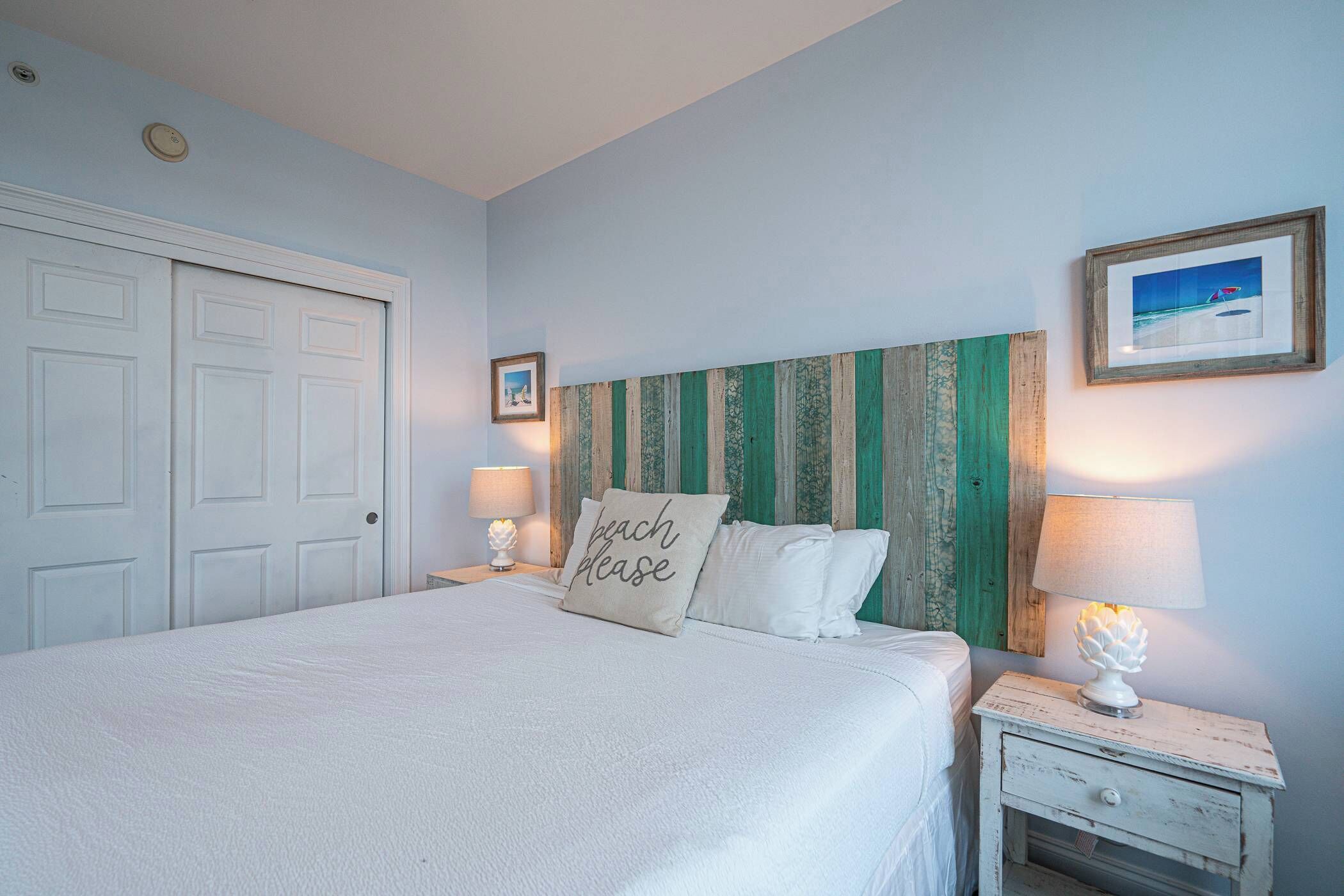 Une chambre à coucher comprenant un lit, deux tables de chevet, une commode, une porte et un mur orné d’un cadre de tableau.