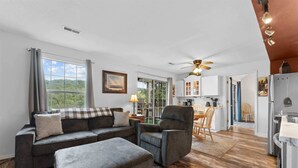 Ein Wohnzimmer mit Sofa, Sessel und Hocker, ein Essbereich mit Stühlen, eine Küche mit Schränken und Geräten sowie ein Deckenventilator.