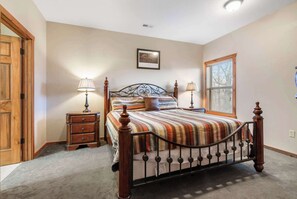 A bedroom with a bed, two bedside tables, a wooden dresser, a window with blinds, and a door.