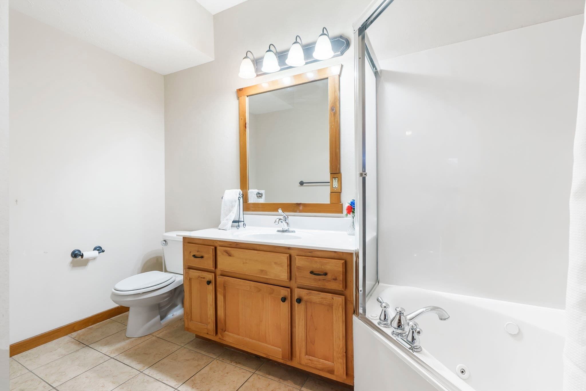 A bathroom with a wooden vanity, a white toilet, a large mirror with lights, and a bathtub.