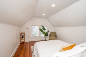 A bedroom with a sloped ceiling, a bed with white bedding, a chair, a plant, and a wooden floor.