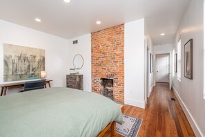 A bedroom with a brick fireplace, a bed with a green bedspread, a desk with a lamp, and a large abstract painting on the wall.