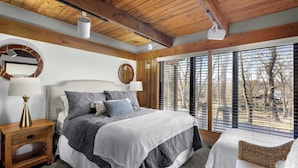 Un dormitorio con una cama grande, vigas de madera en el techo, una ventana con persianas y una mesita de noche con lámpara.