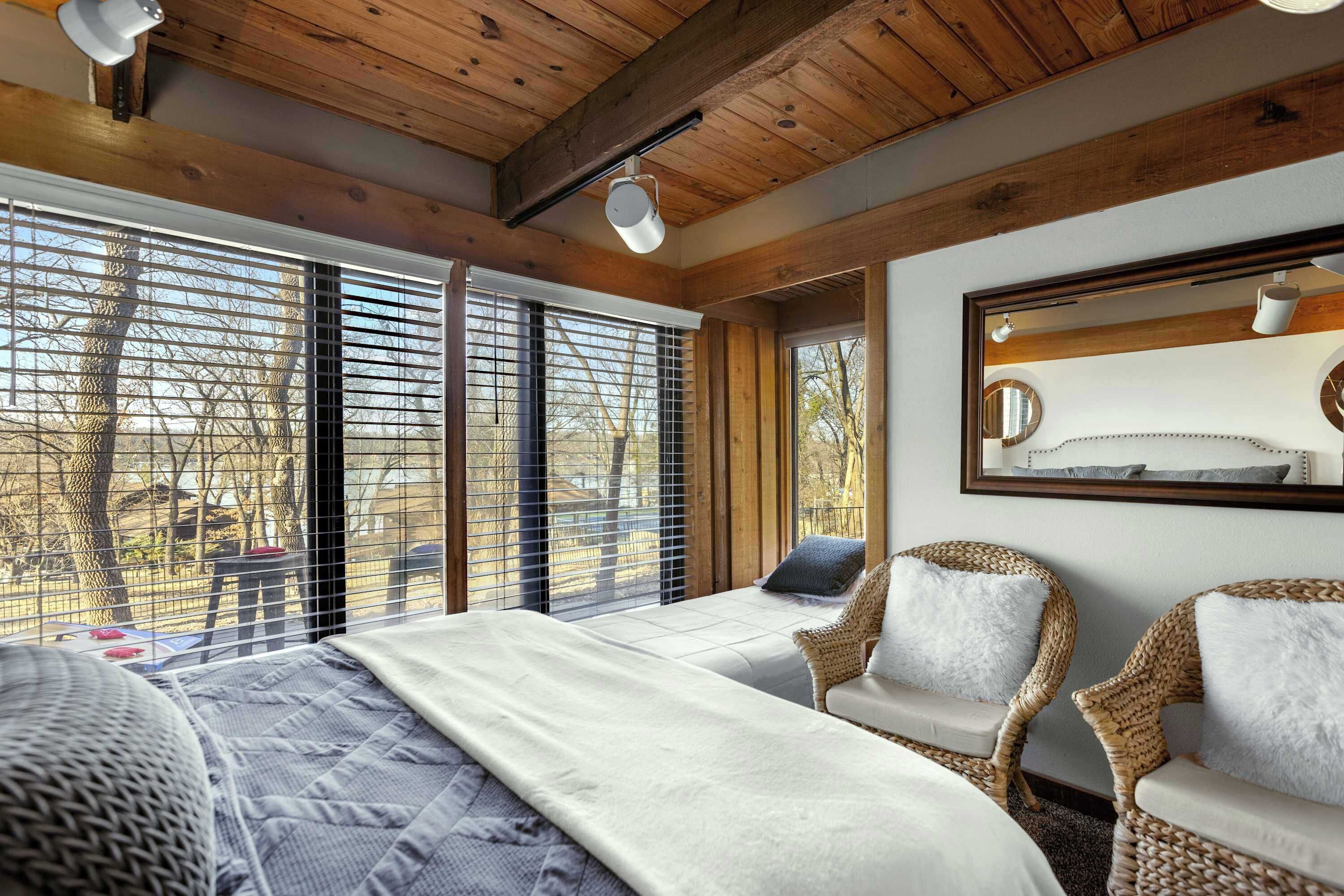 A bedroom with a large bed, two wicker chairs, a wooden ceiling, and a large window with blinds.