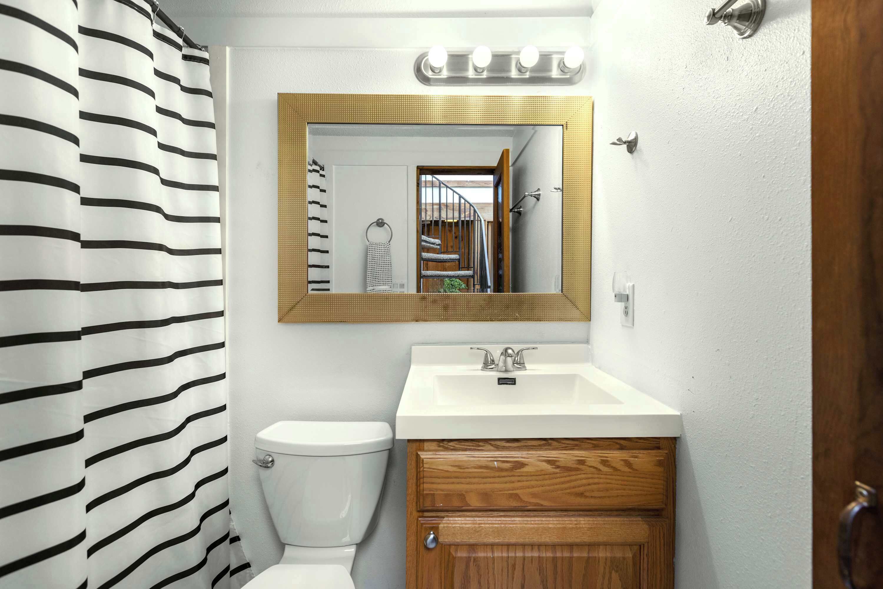 Un baño con cortina de ducha a rayas, un inodoro blanco, un tocador de madera con un espejo sobre el lavabo y un perchero para toallas.