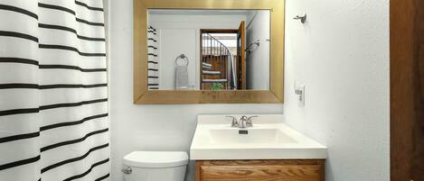Un baño con cortina de ducha a rayas, un inodoro blanco, un tocador de madera con un espejo sobre el lavabo y un perchero para toallas.