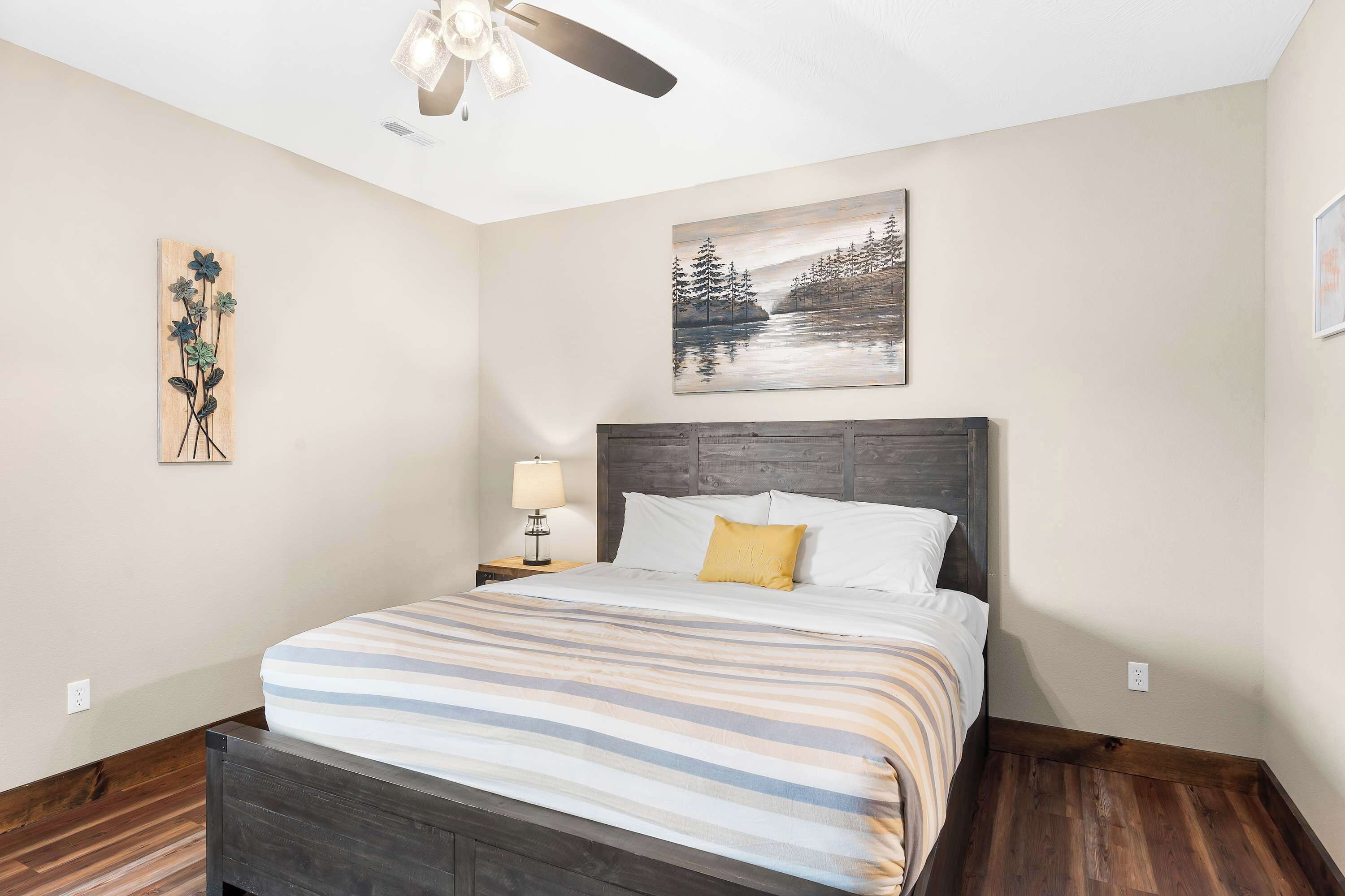 A bedroom with a bed, a nightstand with a lamp, a ceiling fan, and two wall art pieces.