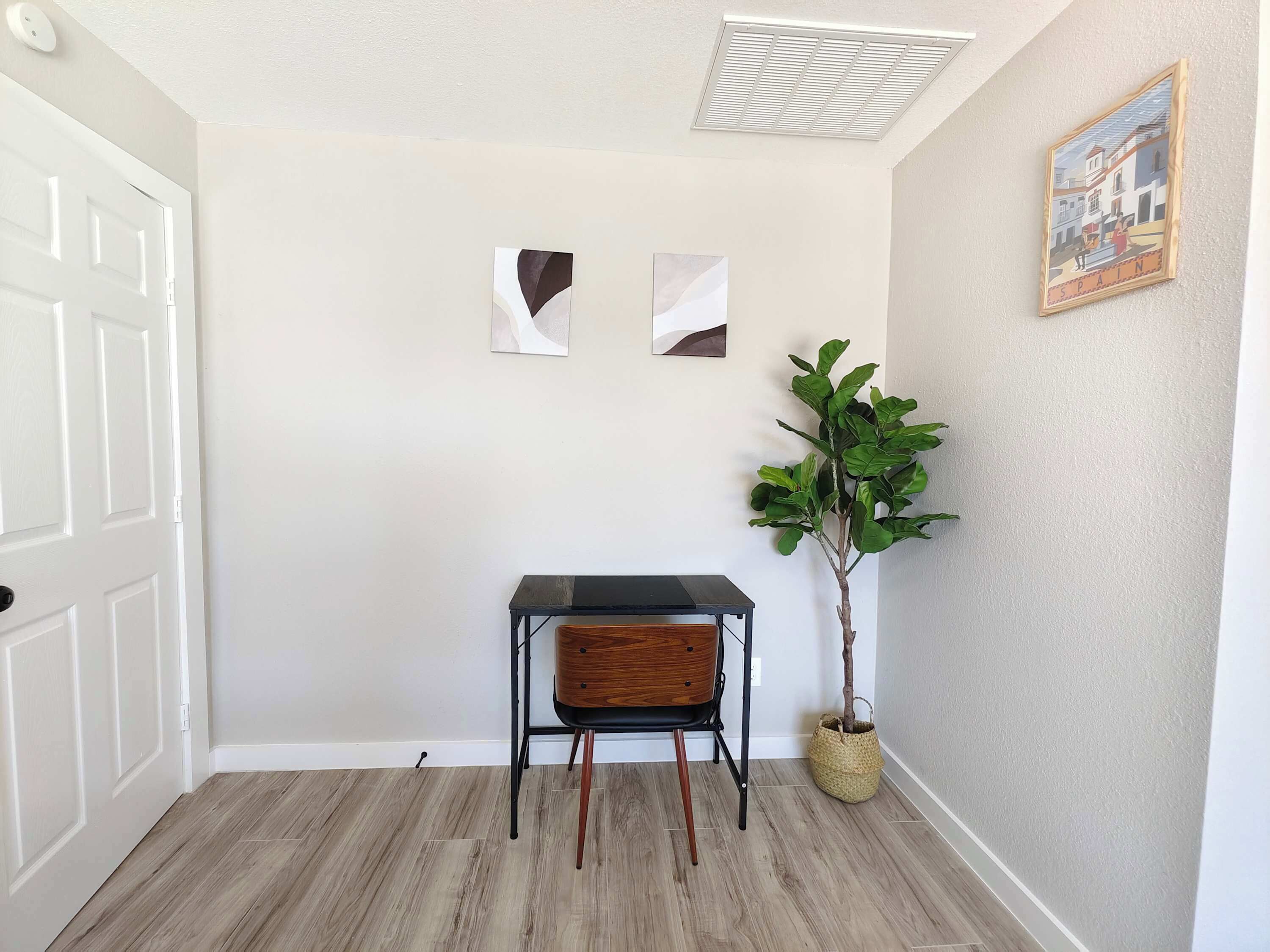 A room with a white door, a small table with a chair, two abstract wall art pieces, a potted plant, and a framed picture on the wall.