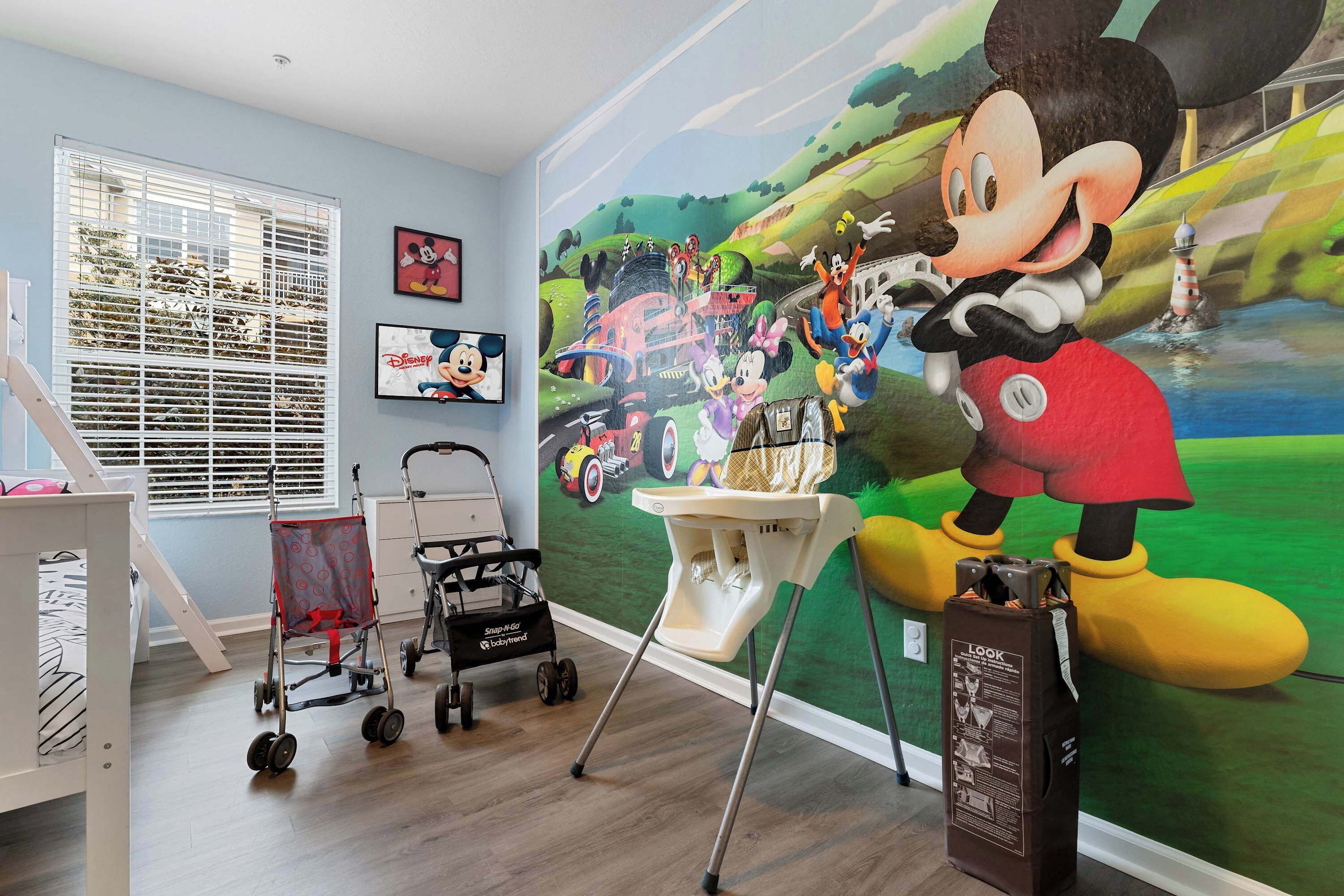 Une pièce décorée d’une grande fresque représentant Mickey Mouse, avec un berceau, une poussette et une chaise haute.