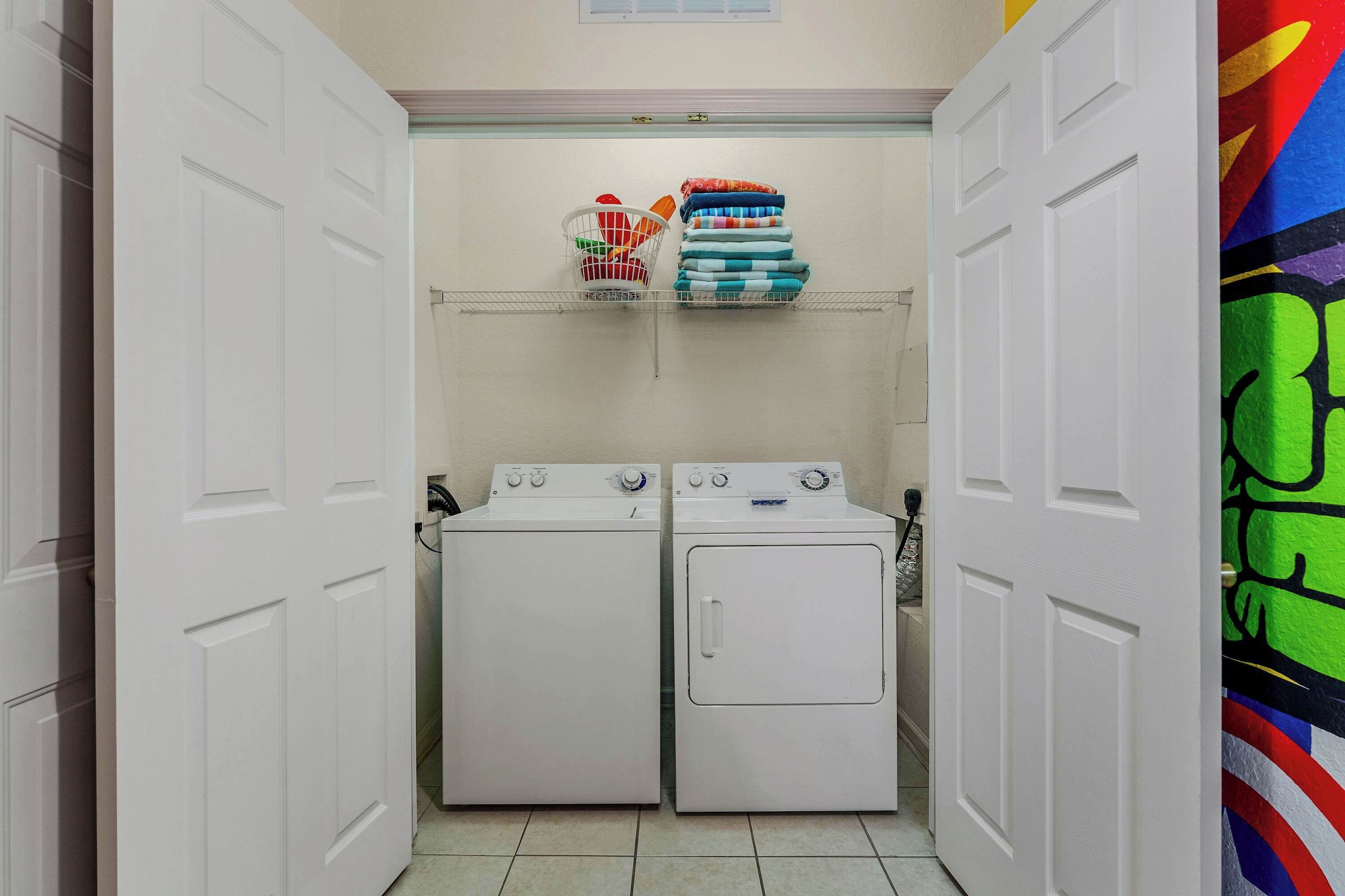A laundry room with two white washing machines, a metal rack with folded clothes, and a colorful wall mural.