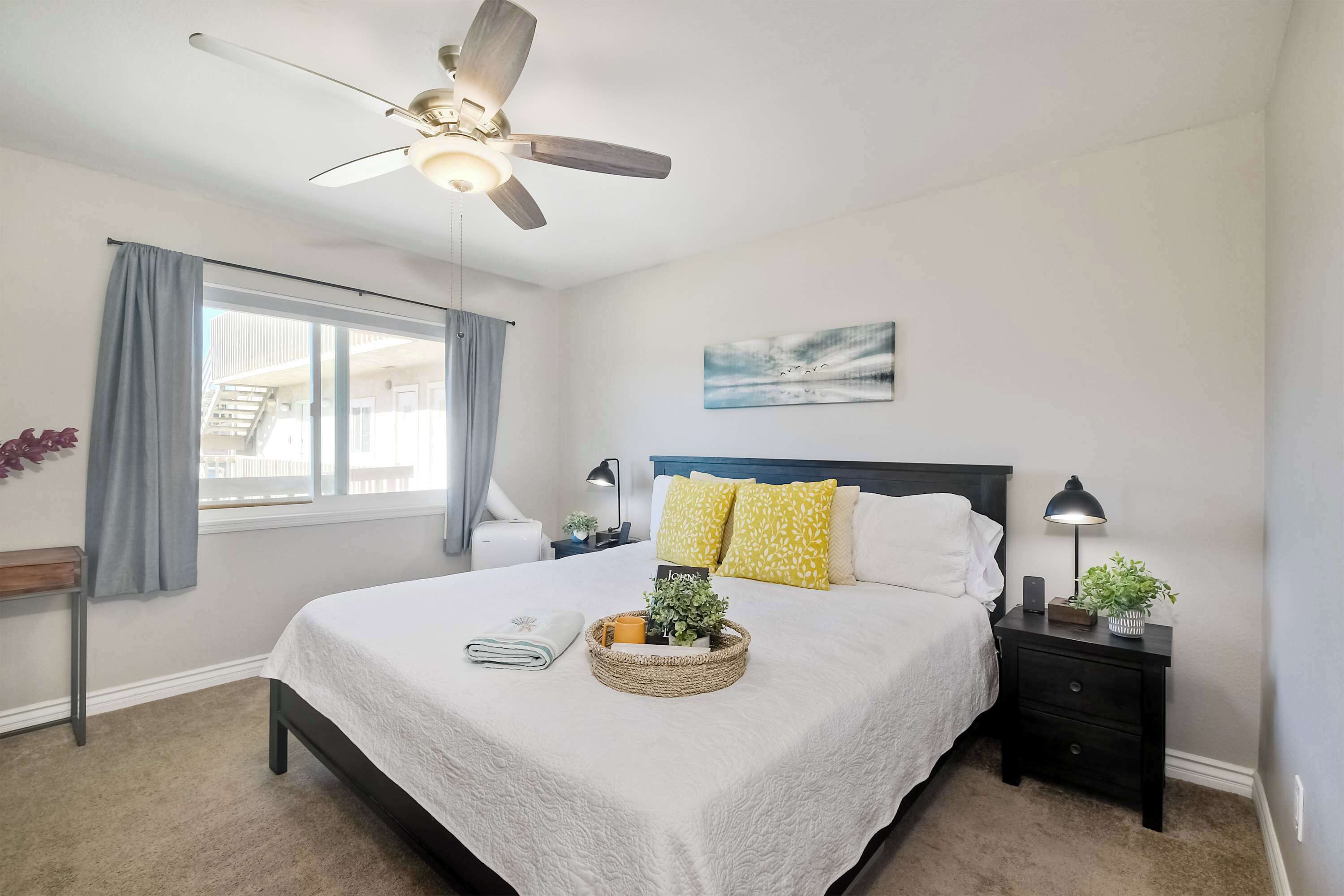 Une chambre à coucher avec un lit, un ventilateur de plafond, une table de chevet, une lampe, une fenêtre avec des rideaux et un tableau au mur.