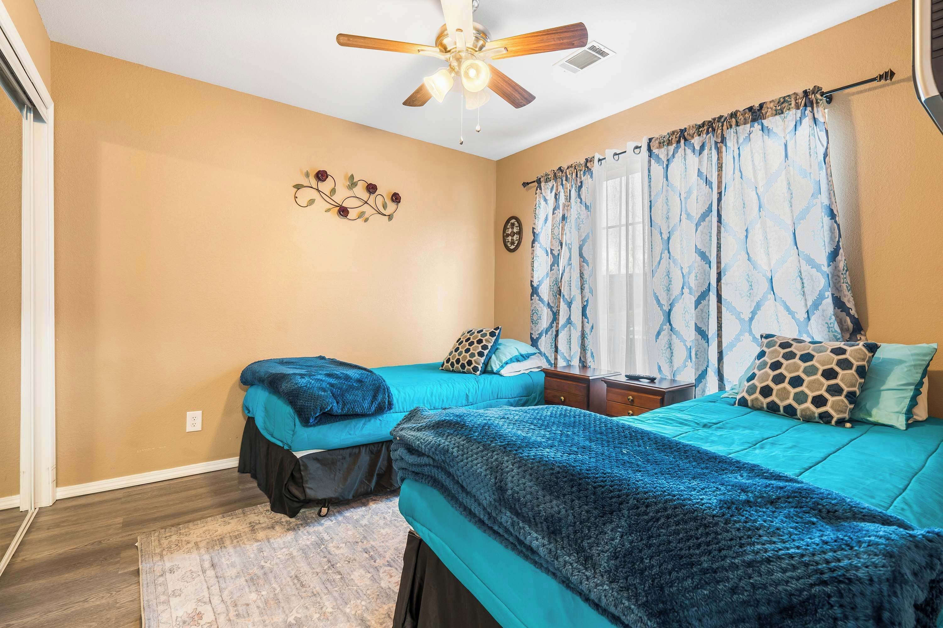 A bedroom with two beds, a ceiling fan, a window with patterned curtains, and a decorative wall piece.