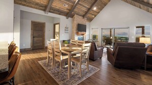 A spacious living room with a high ceiling, wooden flooring, a dining area with a wooden table and chairs, a sofa, and a TV mounted on a brick wall. - Two Pines Endless Views and Attractions! (Branson)
