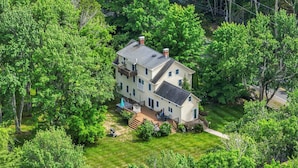 Une maison à deux étages avec un grand jardin à l’avant et une terrasse, entourée d’arbres denses.