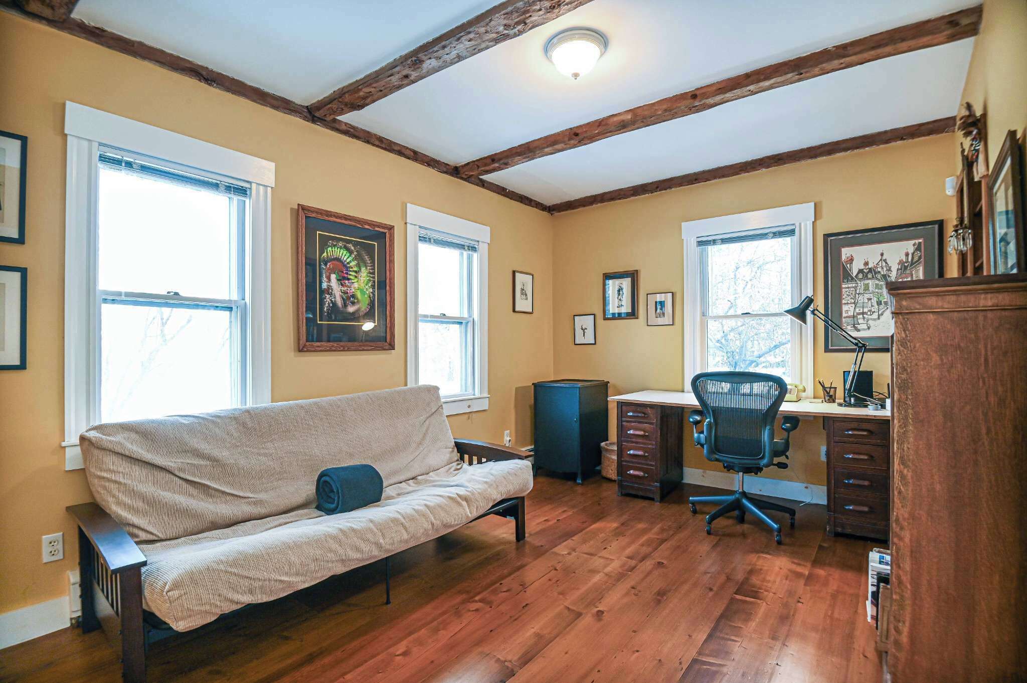 A room with a wooden floor, a sofa, a desk with a chair, and framed pictures on the walls.