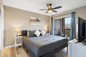 Un dormitorio con cama, lámparas de noche, ventilador de techo, un cuadro en la pared y vistas al exterior a través de una puerta corredera.