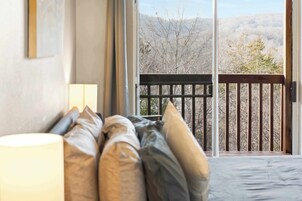 Un dormitorio con un ventanal grande, una cama con muchas almohadas y vistas a árboles y colinas.