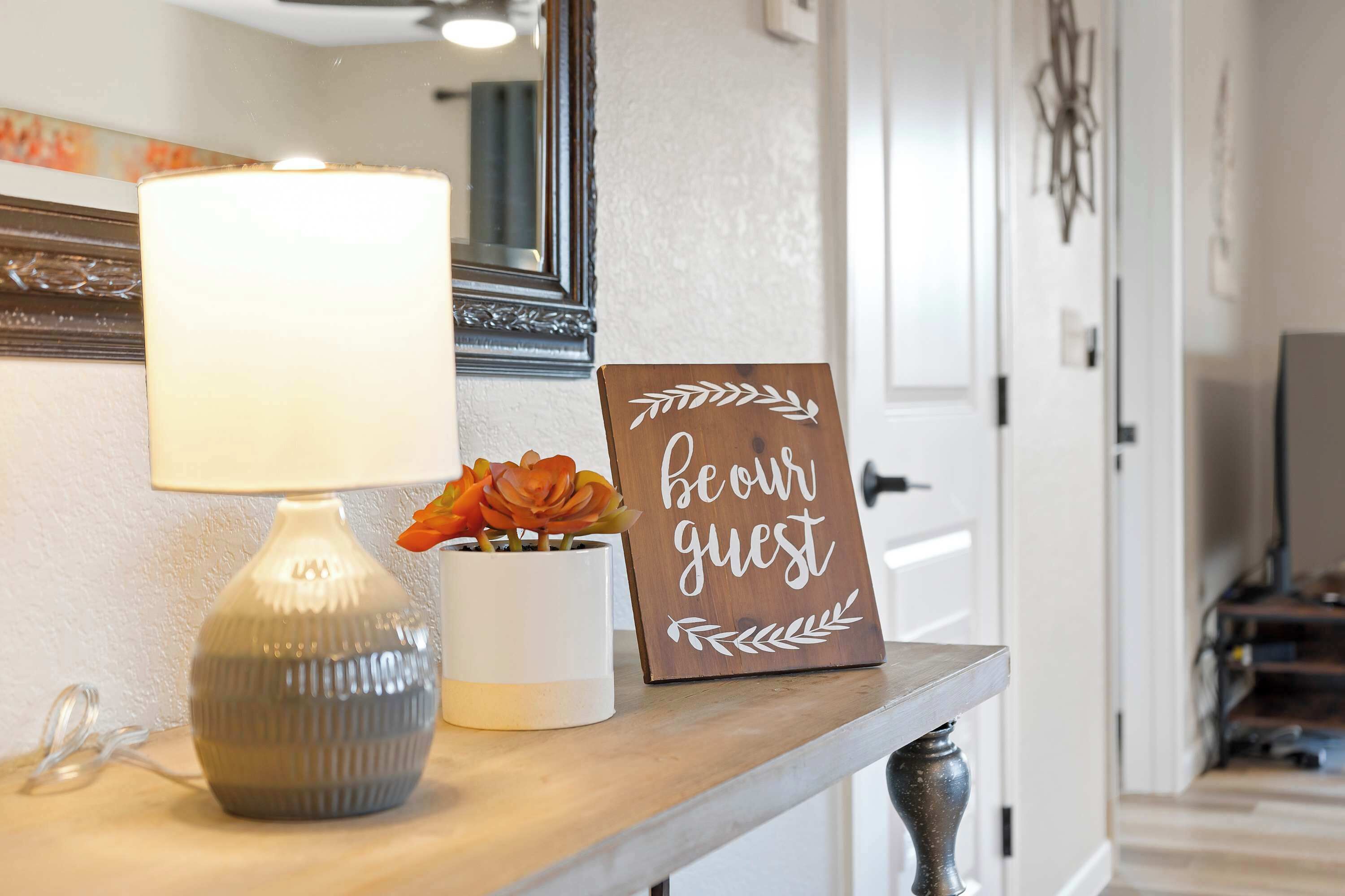 Eine Lampe, ein Schild mit der Aufschrift 'be our guest' und eine Vase mit Blumen auf einem Holzregal.