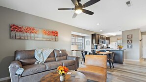 Un saló modern amb un sofà, una butaca i una taula de centre, una zona de menjador amb cadires, i una cuina al fons.