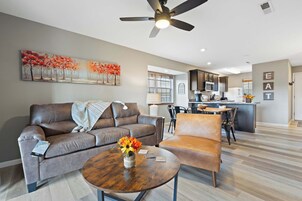 Una sala moderna con sofá, sillón y mesa de centro, un comedor con sillas y una cocina al fondo.