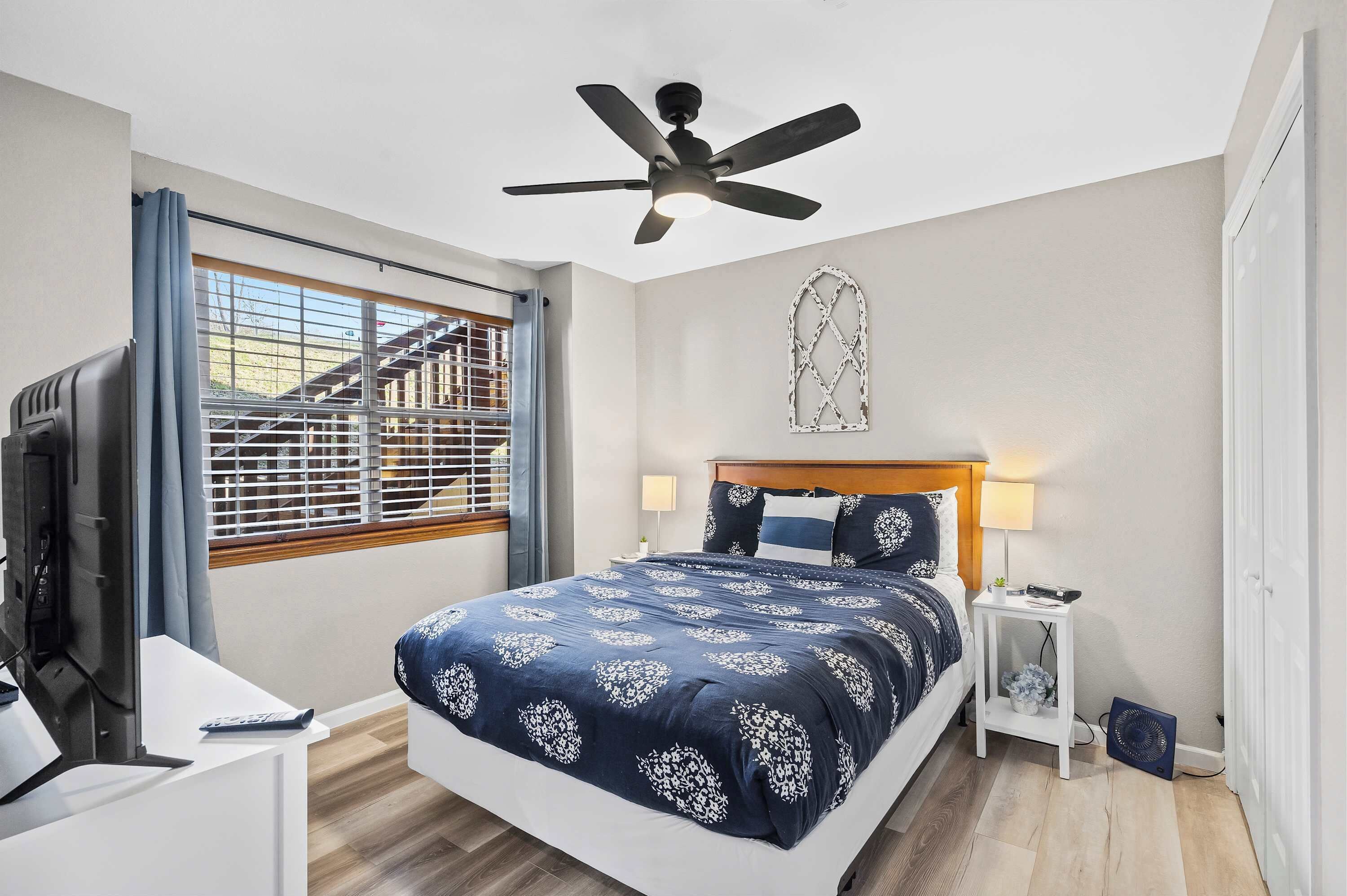 A bedroom with a bed, a ceiling fan, a TV, a desk, and a window with a view of a staircase.