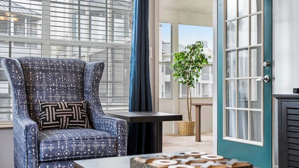 A room with a patterned armchair, a small table with a decorative box, and a view through a window with blinds. - Brookside Haven - Gorgeous, Private Community! (Branson)