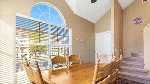 A dining area with a wooden table and chairs, a large window with blinds, and a staircase leading to another floor.