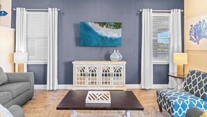 A living room with a dark wooden coffee table, a grey sofa, a patterned armchair, a white cabinet with glass doors, and a large framed picture of a beach scene. - Lafitte's Landing Lakeview Cottage - Lakefront! (Kissimmee)