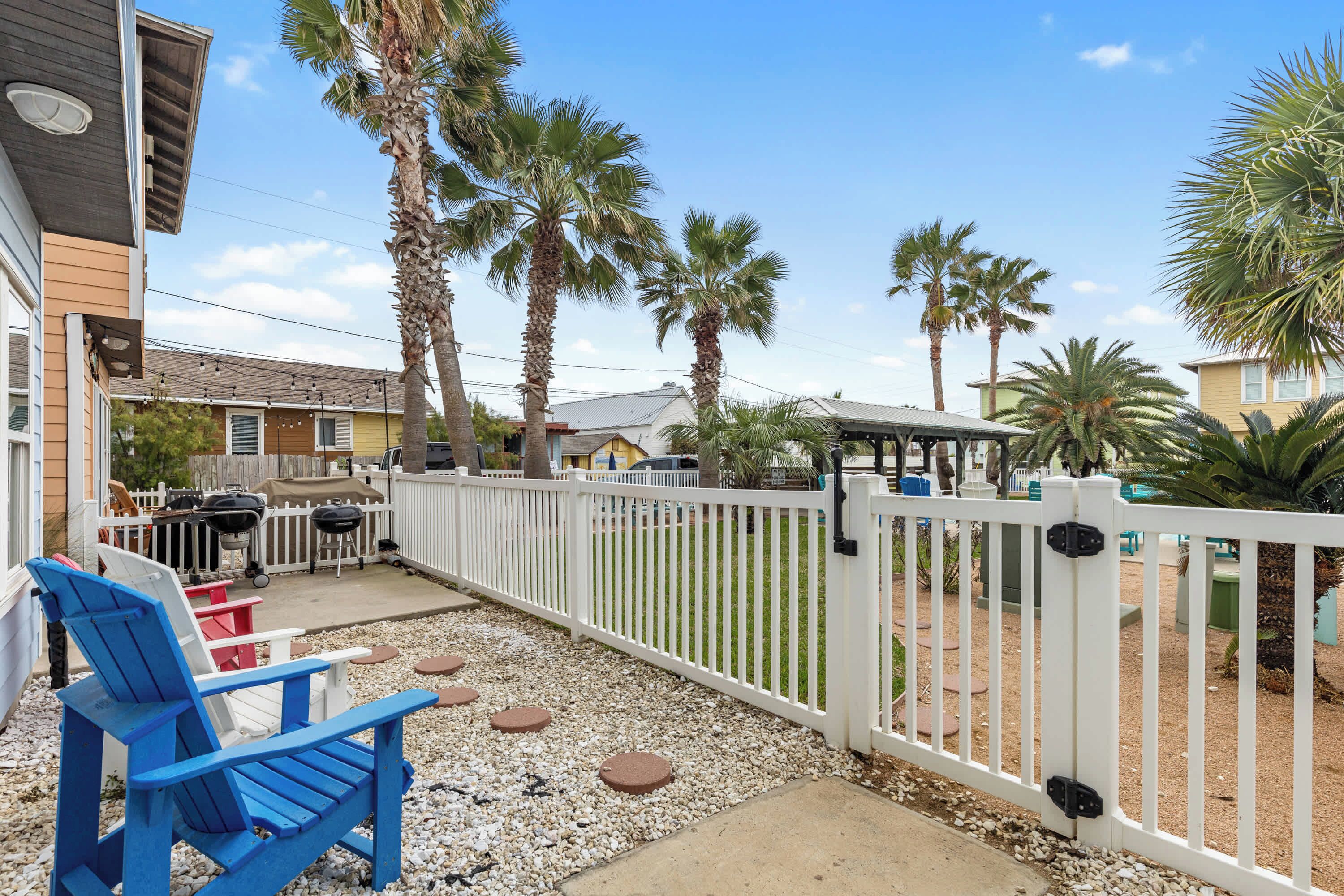 A backyard with a white fence, blue and red Adirondack chairs, palm trees, and a barbecue grill.
