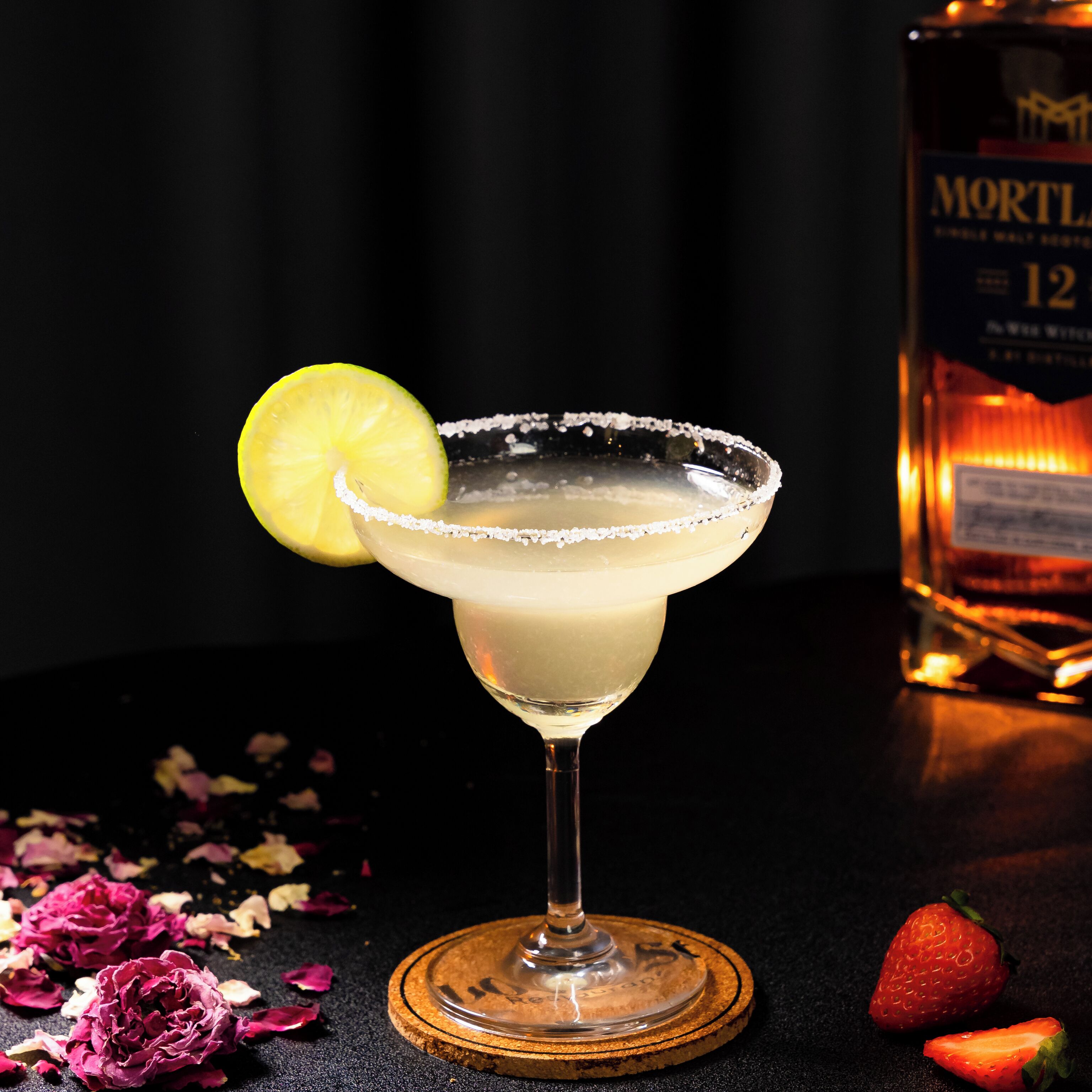 A margarita cocktail in a glass with a salted rim, garnished with a lemon slice, on a wooden coaster. A bottle of Mortl is visible in the background.