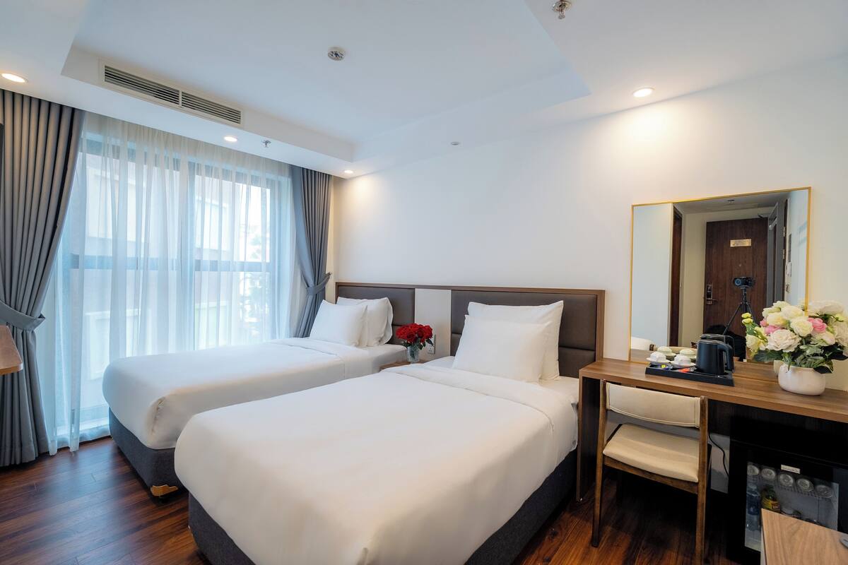 A hotel room with two beds, a desk with a mirror, a chair, and a small table with a vase of flowers.