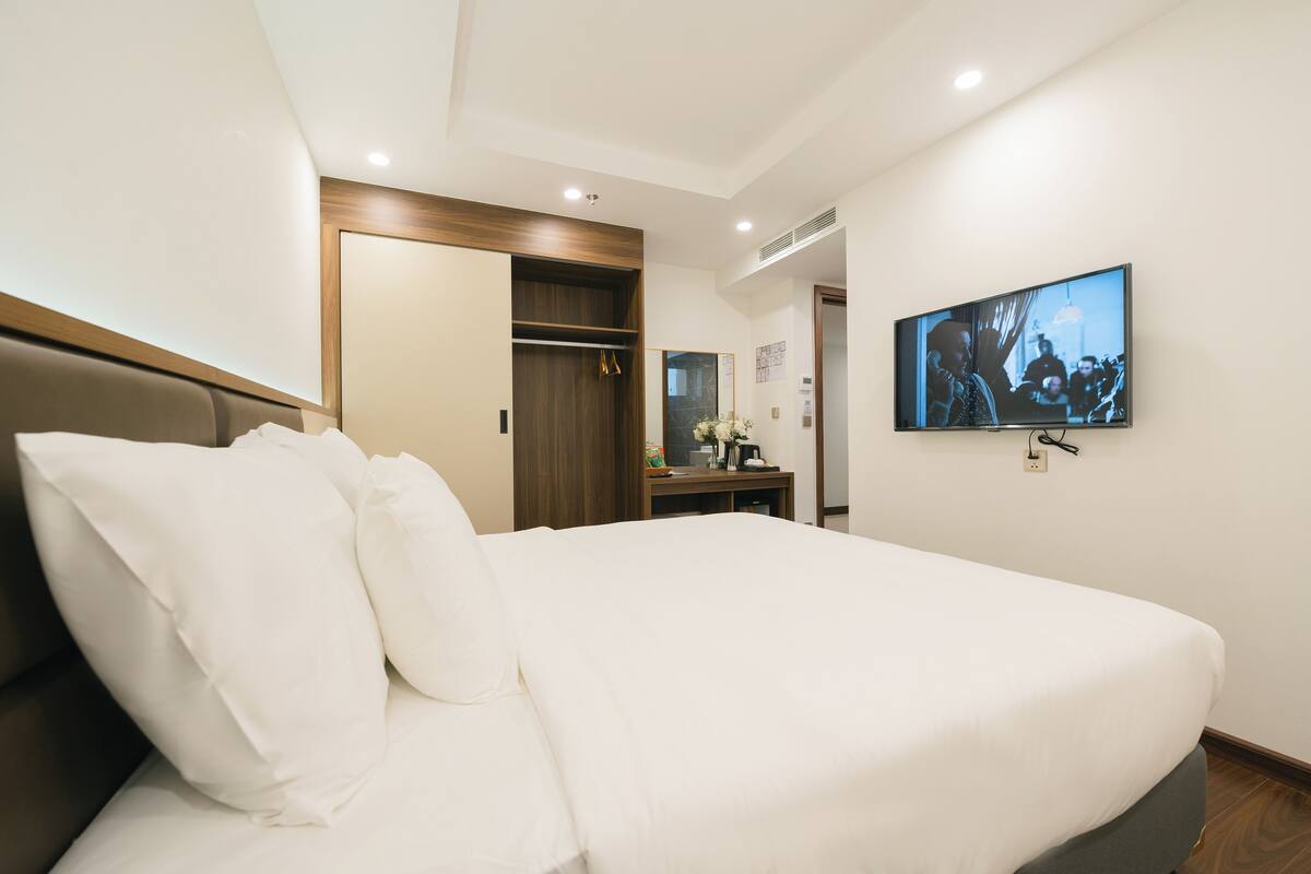 A hotel room with a large bed, a TV mounted on the wall, a wooden wardrobe, and a small table with a vase of flowers.
