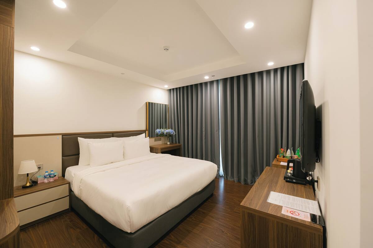 A hotel room with a large bed, wooden headboard, bedside tables with lamps, a desk with a flat-screen TV, and a curtain-covered window.