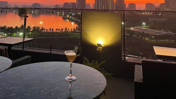 A sunset view from a terrace with a table and a drink, overlooking a cityscape and a body of water. - Love St Hotel (Hanoi)
