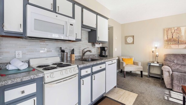 A compact kitchen with white and gray cabinets, a built-in microwave, and a stove. There is a small dining area with a chair and a side table. - Windsor Cottage Fall Creek Beauty by Table Rock! (Branson)